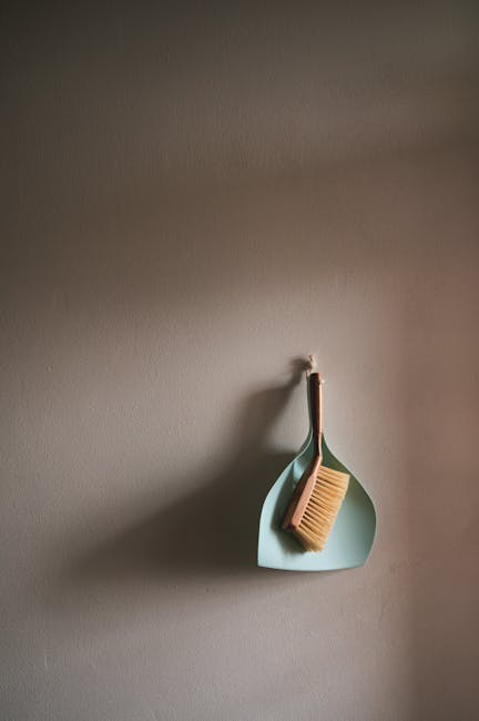 A wall-mounted hook holding a dustpan and broom with wooden handles and natural bristles, both hanging on a plain, light-colored wall. The dustpan is made of light blue plastic, positioned beneath the